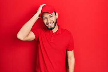 Hispanic man with beard wearing delivery uniform and cap smiling confident touching hair with hand up gesture, posing attractive and fashionable