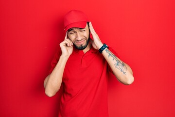 Hispanic man with beard wearing delivery uniform and cap covering ears with fingers with annoyed expression for the noise of loud music. deaf concept.