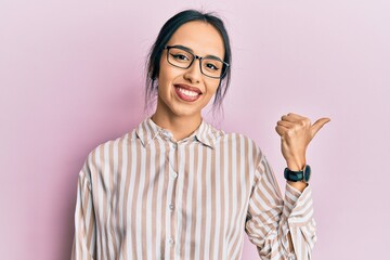 Young hispanic girl wearing casual clothes and glasses pointing thumb up to the side smiling happy...