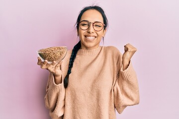 Young hispanic girl holding lentils bowl screaming proud, celebrating victory and success very...