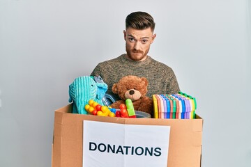 Young redhead man holding donation box with toys skeptic and nervous, frowning upset because of...