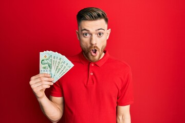 Young redhead man holding 20 thai baht banknotes scared and amazed with open mouth for surprise, disbelief face