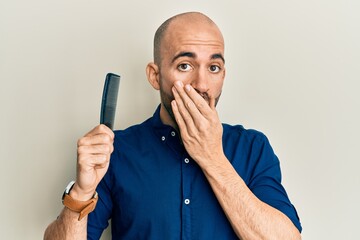 Young hispanic man holding comb loosing hair covering mouth with hand, shocked and afraid for...