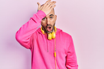 Young bald man wearing gym clothes and using headphones surprised with hand on head for mistake,...