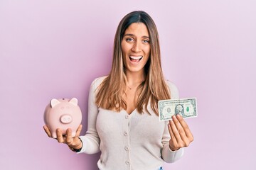 Brunette young woman holding one dollar banknote and piggy bank celebrating crazy and amazed for success with open eyes screaming excited.