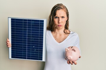 Young blonde woman holding photovoltaic solar panel and piggy bank skeptic and nervous, frowning...