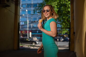 A happy woman in a turquoise dress stands in an arched passage and eats a waffle cone on a warm summer day. Beautiful blonde in sunglasses enjoys ice cream while walking. Summer dessert.
