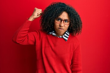 Fototapeta na wymiar Beautiful african american woman with afro hair wearing sweater and glasses angry and mad raising fist frustrated and furious while shouting with anger. rage and aggressive concept.