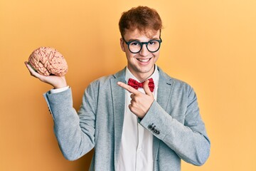 Young caucasian man wearing glasses holding brain smiling happy pointing with hand and finger