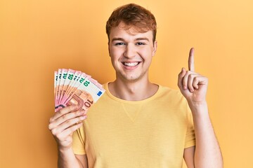 Young caucasian man holding bunch of 10 euro banknotes smiling with an idea or question pointing finger with happy face, number one