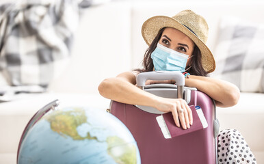 Woman in summer hat and face mask leans against the suitcase holding flight ticket and a passport...