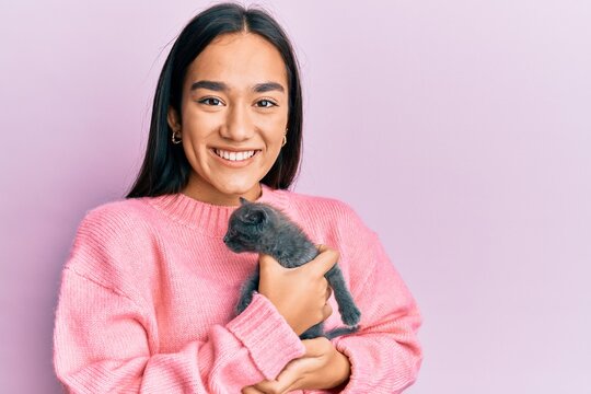 Young hispanic girl smiling happy holding cute cat over isolated pink background.