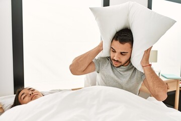 Man covering ears with pillow irritated for snore lying in bed at home.