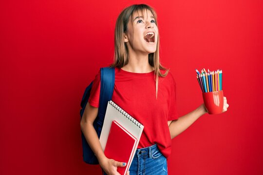 Teenager caucasian girl holding art notebooks and colored pencils angry and mad screaming frustrated and furious, shouting with anger looking up.