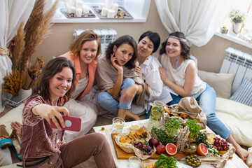 Group of happy young female friends posing taking selfie use smartphone at hen-party in hippie style