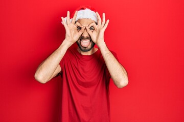 Young hispanic man wearing christmas hat doing ok gesture like binoculars sticking tongue out, eyes looking through fingers. crazy expression.