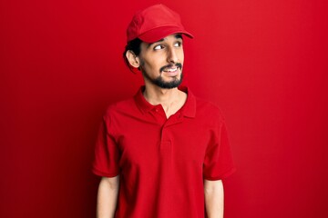 Young hispanic man wearing delivery uniform and cap looking away to side with smile on face, natural expression. laughing confident.