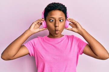 Young african american girl listening to music using headphones making fish face with mouth and squinting eyes, crazy and comical.