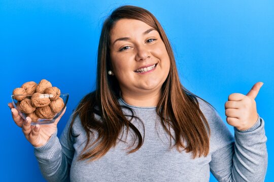 Beautiful brunette plus size woman holding bowl with walnuts pointing thumb up to the side smiling happy with open mouth