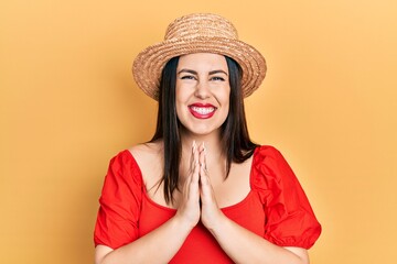 Young hispanic woman wearing summer hat praying with hands together asking for forgiveness smiling confident.
