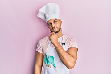 Young arab man wearing professional cook apron and hat thinking concentrated about doubt with finger on chin and looking up wondering