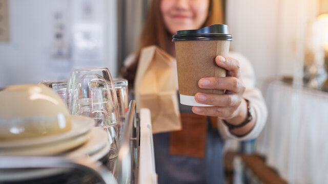 Closeup image of a waitress holding and serving paper cup of coffee and takeaway food in paper bag to customer in a shop
