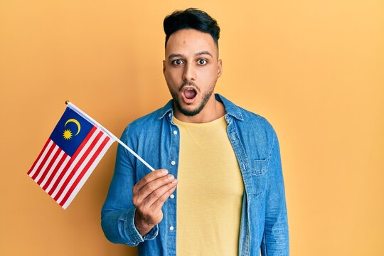 Young Arab Man Holding Malaysia Flag Scared And Amazed With Open Mouth For Surprise, Disbelief Face