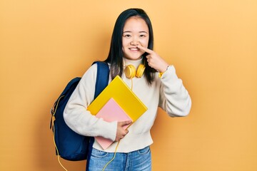 Young chinese girl holding student backpack and books pointing with hand finger to face and nose, smiling cheerful. beauty concept