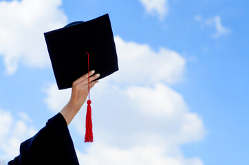 Closed up female university student's hand holding black graduate hat over blue sky background