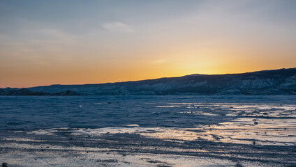 Fototapeta na wymiar Sunset over a frozen lake. The sky above the mountain range is colored orange. On the ice there are patches of snow and reflections of the setting sun. Baikal