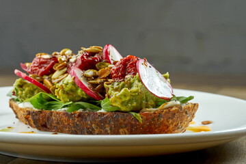Avocado toast with guacamole, radish, sun-dried tomatoes and seeds on rye bread, served in a white plate on a wooden background. Vegetarian food