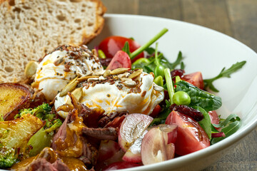 Appetizing and hearty breakfast with poached egg, potatoes, bacon, grapes, mix salad and rye bread in a white bowl on a wooden background
