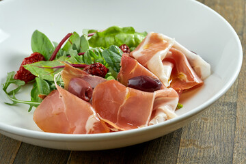Mix salad with jamon crudo, olives and sun-dried tomatoes in a white bowl on a wooden background.