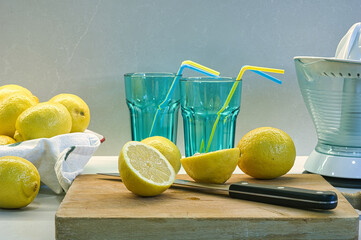 Limones cortados sobre una tabla de cocina, vasos azules, exprimidor blanco y fondo gris.