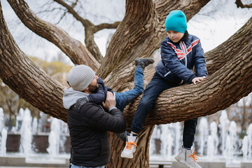 children sitting on big tree in park. A father helping his cute little son coming down . Image with selective focus.