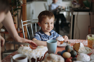 cute little boy eating his easter cake and drinking tea sitting at the table covered with tablecloth in living room. Woman's hand cutting a piece of cake on foreground. Image with selective focus 