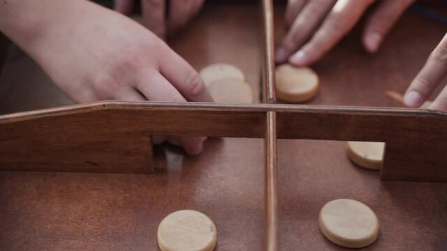 Four caucasian hands are very active in playing a wooden pucket. Wooden chips quickly fall into the holes. Close-up view of the center of table.