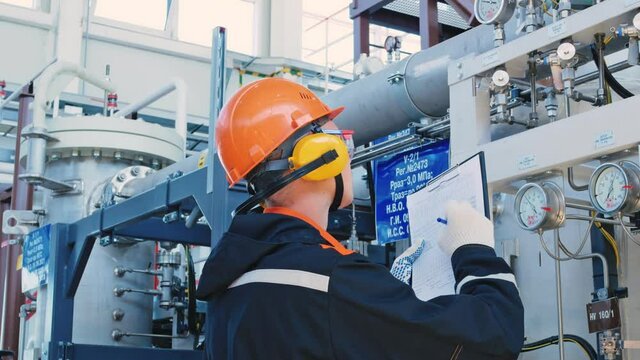 A worker in a helmet, goggles and headphones, writes data on a sheet of paper, monitors the pressure and temperature readings. Work in industrial. Manufacturing industry.