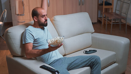 Concentrated man watching entertainment movie while making face expression, holding popcorn bowl in hands. Freelancer in pajamas sitting on sofa looking at entertainment shows late at night in kitchen