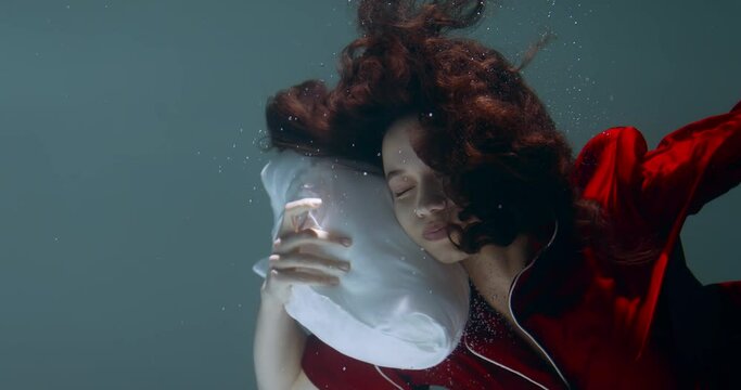 Cinematic close-up, young beautiful mixed race woman in red pyjamas sinking under water, head on pillow slow motion.
