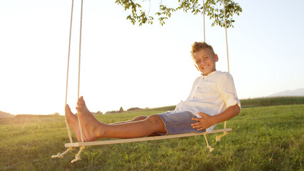 CLOSE UP Young boy relaxing on a wooden swing at sunset, posing for the camera. - obrazy, fototapety, plakaty