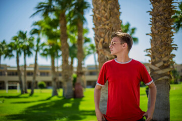 cute young teenage boy posing and having fun on the beach under palm trees in red T-shirt . Trendy boy posing. Funny and positive man in sunglasses