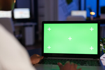 Back view of black business woman using laptop with green screen, green mockup, chroma key desktop sitting at desk in start up business office overworking. Freelancer watching at isolated display.