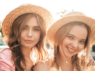 Two young beautiful smiling hipster woman in trendy summer sundress. Sexy carefree women posing on the street background in hats. Positive models having fun on embankment at sunset