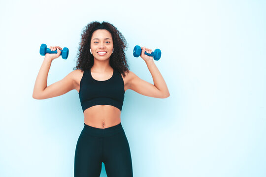 Fitness smiling black woman in sports clothing with afro curls hairstyle.She wearing sportswear. Young beautiful model with perfect tanned body.Female holding dumbbells in studio near light blue wall © halayalex