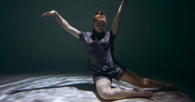 Young beautiful redhead woman wearing pyjamas falling down deep under water looking at camera in dark pool slow motion.