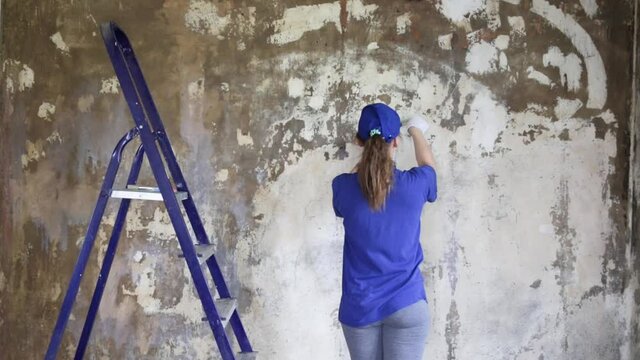 A young woman in a blue t-shirt and a baseball cap makes repairs in the apartment. Apply a primer to the walls