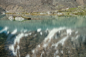 Rock in turquoise mountain lake. Snowy mountain reflected in azure clear water of glacial lake. Beautiful sunny background with snow-white glacier reflection in green water surface of mountain lake.