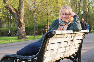 Portrait of beautiful middle-aged woman in the park on bench. Woman with blond hair, in jeans and sweater, is resting in city park.