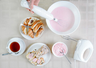 woman is preparing pink cream with a blender in a white plate on a table with pastries, desserts...